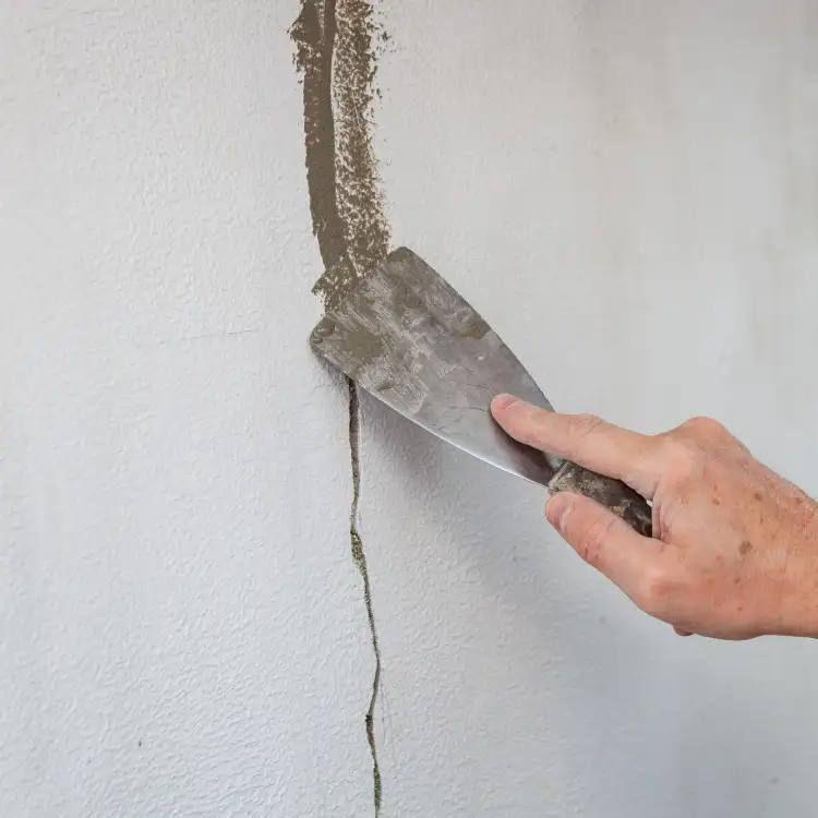 Ręka rozprowadza masę szpachlową na pęknięciu ściany przy pomocy metalowej szpachelki