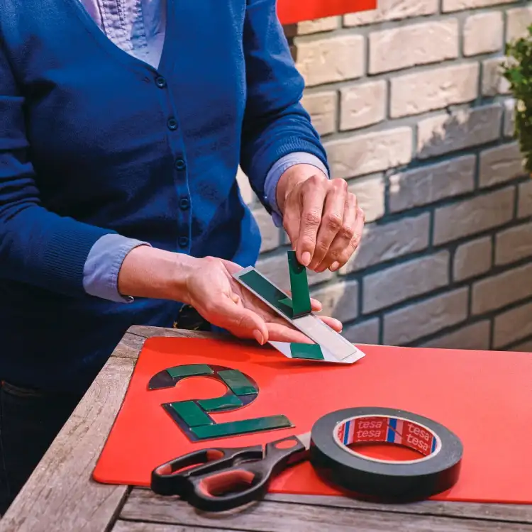Na stole widoczna rolka piankowej taśmy Tesa oraz wycinane cyfry przygotowywane do montażu