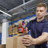 Mężczyzna sprawnie owija karton taśmą pakową za pomocą metalowej zaklejarki Tesa
