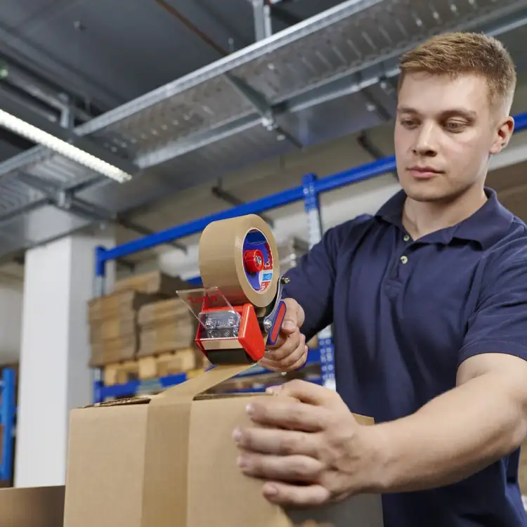 Mężczyzna sprawnie owija karton taśmą pakową za pomocą metalowej zaklejarki Tesa