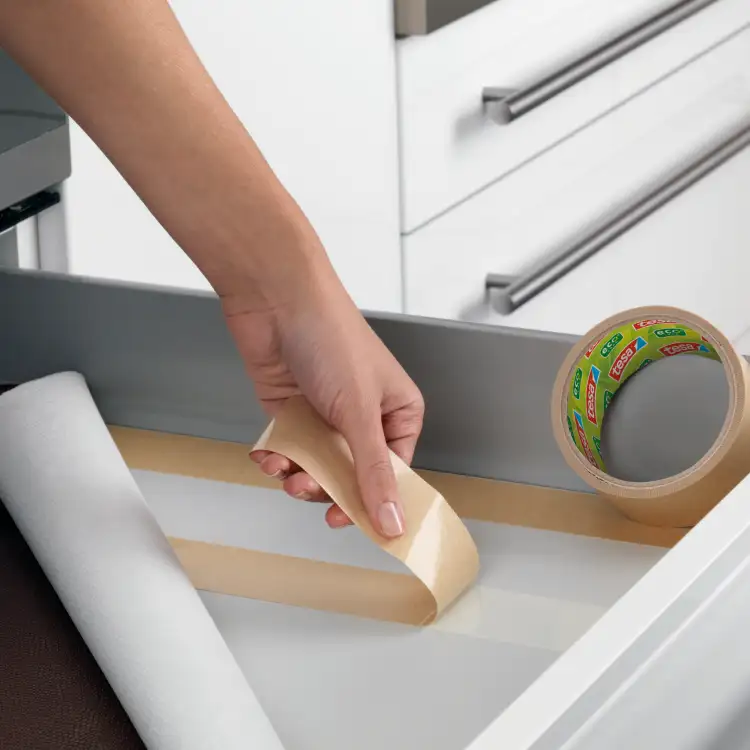 Ręka odkleja papier ochronny taśmy Tesa Eco podczas montażu wykładziny w szufladzie kuchennej