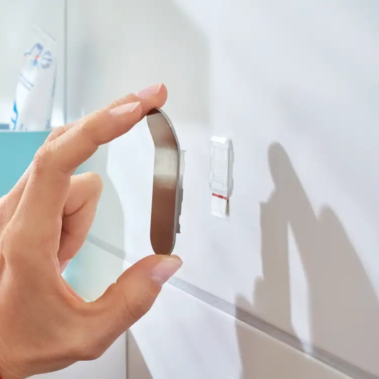Zbliżenie na montaż haczyka łazienkowego Tesa Powerstrips na gładkiej, jasnej płytce ściennej