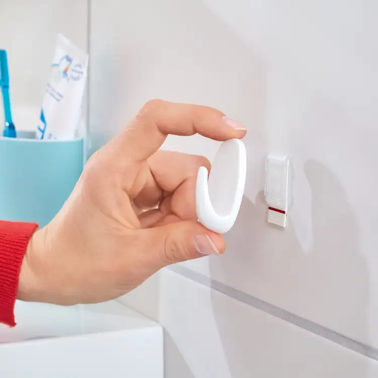 Osoba zbliża biały haczyk łazienkowy Tesa Powerstrips do samoprzylepnej bazy na płytce