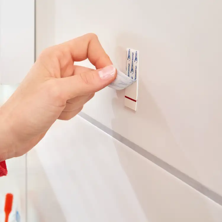 Zbliżenie na montaż samoprzylepnego haczyka Tesa Powerstrips na białych płytkach łazienkowych