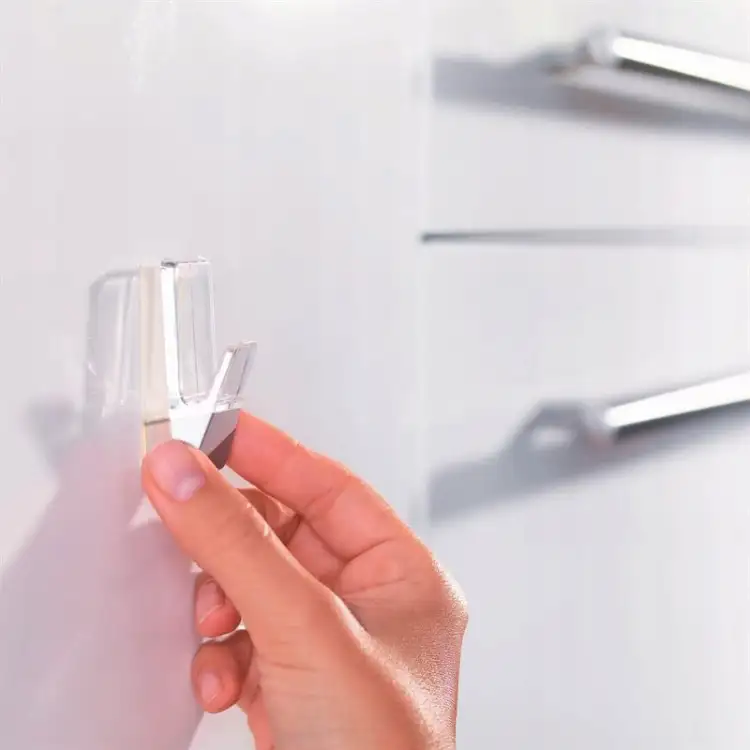 Delikatna dłoń zbliża się do przezroczystego haczyka łazienkowego Tesa Powerstrips na jasnej szafce