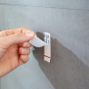 Palce odklejają zabezpieczenie taśmy samoprzylepnej haczyka łazienkowego Tesa Powerstrips na szarej płytce