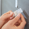 Dłonie odklejają pasek samoprzylepny Tesa Powerstrips przed zamocowaniem na szarej ścianie