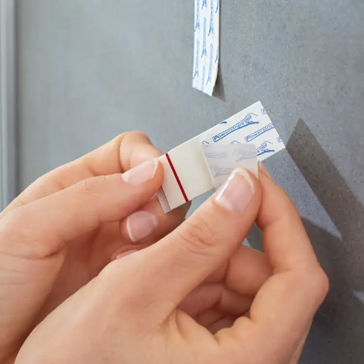 Dłonie odklejają pasek samoprzylepny Tesa Powerstrips przed zamocowaniem na szarej ścianie