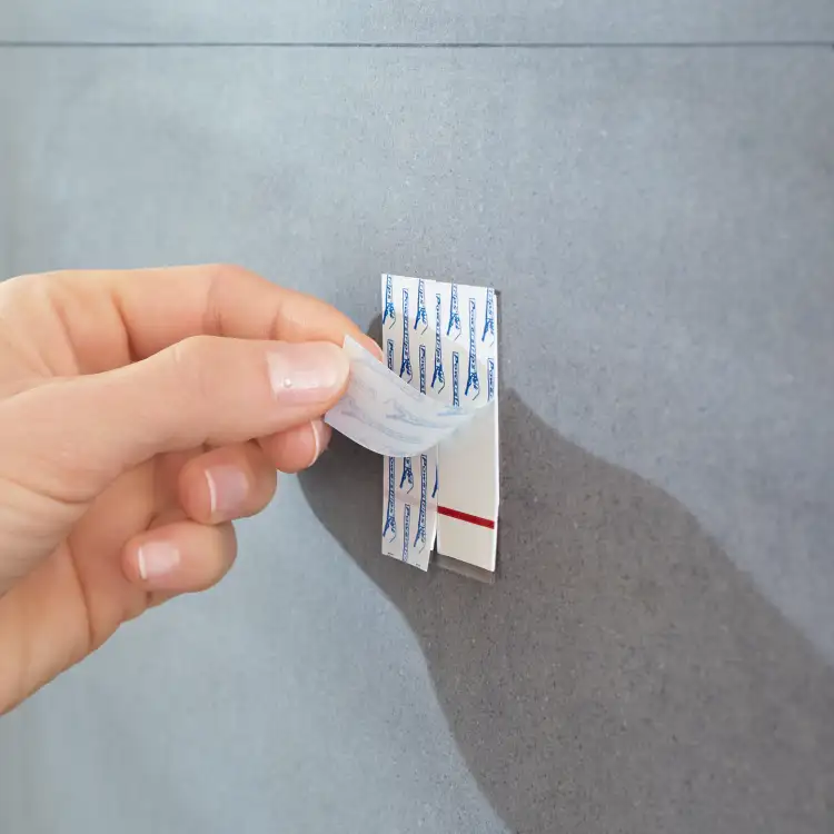 Palce odsłaniają warstwę ochronną samoprzylepnej taśmy Tesa Powerstrips na metalowym haczyku łazienkowym