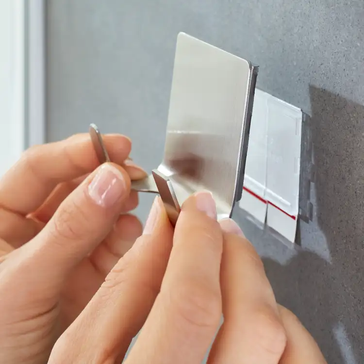 Srebrny metalowy haczyk łazienkowy Tesa Powerstrips przygotowany do montażu na gładkiej ścianie