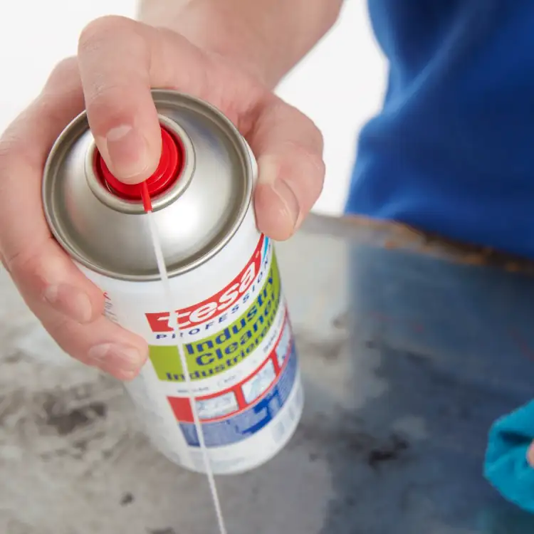 Ręka aplikuje preparat Tesa w sprayu na zabrudzoną powierzchnię metalowego blatu