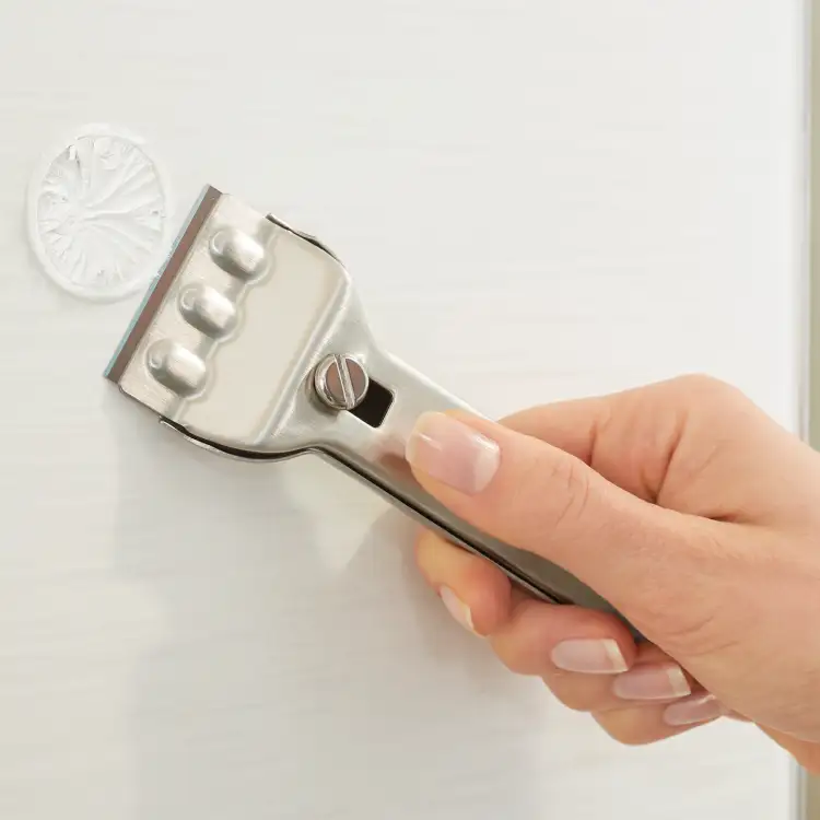 Metalowy skrobak usuwa okrągłą naklejkę montażową Tesa z białej płytki łazienkowej