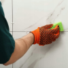 Ręka w pomarańczowej rękawicy z gąbką dokładnie czyści fugi między płytkami na ścianie
